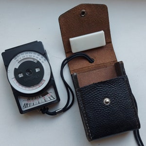 Puede incluir: Un fotómetro blanco y negro con una funda de cuero. El fotómetro tiene un dial circular con la palabra "EDUXFAB" impresa en él. La funda es marrón por dentro y negra por fuera.