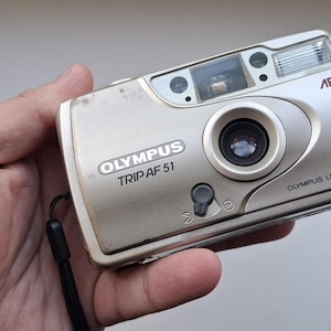 Könnte beinhalten: Silberne Olympus Trip AF 51 Kamera mit 28mm Objektiv. Die Kamera hat einen eingebauten Blitz und der Text "OLYMPUS TRIP AF 51" ist auf der Vorderseite aufgedruckt. Die Kamera wird in einer Hand gehalten.