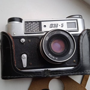 May include: A silver and black 35mm film camera with a leather case. The camera has a lens with the text "N-61/A 2.8/53 9428203" printed on it. The camera also has the text "ФЗО-5" printed on it.