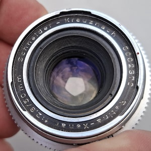 May include: A silver and black camera lens with the text Schneider-Kreuznach Retina-Xenar f:2.8/50mm and the serial number 5062370 engraved on the front ring. The lens features a circular glass element with a visible pentagonal aperture opening.