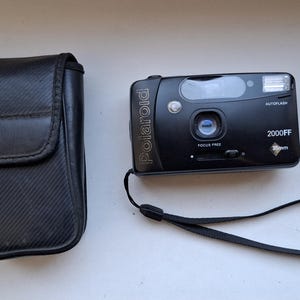 Op de afbeelding: Een zwarte Polaroid 2000FF camera met een zwarte leren hoes en een zwarte band. De camera heeft een ingebouwde flitser en een lensdop.