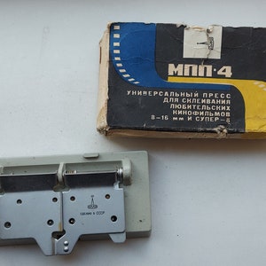 May include: A gray metal film splicing press labeled "MΠΠ-4" in black lettering. The press is labeled "УНИВЕРСАЛЬНЫЙ ПРЕСС" in black lettering, and "ДЛЯ СКЛЕИВАНИЯ ЛЮБИТЕЛЬСКИХ КИНОФИЛЬМОВ 8-16 мм И СУПЕР-8" in black lettering. The press is sitting on a white surface. The press is in a box with a blue and yellow design. The box is labeled "MΠΠ-4" in black lettering. The box is labeled "УНИВЕРСАЛЬНЫЙ ПРЕСС" in black lettering, and "ДЛЯ СКЛЕИВАНИЯ ЛЮБИТЕЛЬСКИХ КИНОФИЛЬМОВ 8-16 мм И СУПЕР-8" in black lettering.