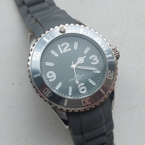 May include: A silver-toned wristwatch with a black face and white markings. The watch has a gray silicone band and a black bezel with white numbers.
