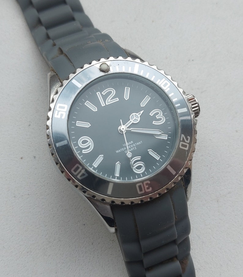 May include: A silver-toned wristwatch with a black face and white markings. The watch has a gray silicone band and a black bezel with white numbers.