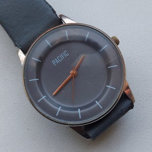 Puede incluir: Un reloj de pulsera de cuero negro con caja plateada y esfera negra. La esfera del reloj tiene marcas blancas y la palabra "PACIFIC" impresa en blanco.