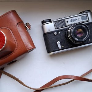 May include: A vintage silver camera with a black leather case and a brown leather strap. The camera has a lens with a silver rim and a black dial that reads "030-5".