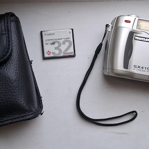 Könnte beinhalten: Eine silberne Digitalkamera mit einer schwarzen Ledertasche und einer Speicherkarte. Die Kamera hat ein Zoomobjektiv und einen eingebauten Blitz. Die Kamera ist mit "Traveler SX410z 4.1 Mega Pixels" beschriftet.
