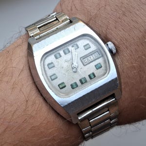 Op de afbeelding: Een zilverkleurig polshorloge met een rechthoekige kast en een metalen schakelarmband. De gebroken witte wijzerplaat heeft donkere rechthoekige uurmarkeringen, zilveren wijzers en een datumvenster met de cyrillische tekst СБТ 8 aan de rechterkant.