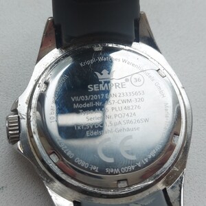 May include: The back of a silver watch with a black band. The watch face has the text "SEMPRE" and "10 bar wasserdicht" printed on it. The watch also has the text "VII/03/2017 EAN 23335653 Modell-Nr. AS7-CWM-320 Type ALS5 PLU 48276 Serier Nr. PO7424 1x1,5V DC 1,5 &mu;&Alpha; SR626SW Edelstahl-Geh&auml;use" printed on it.