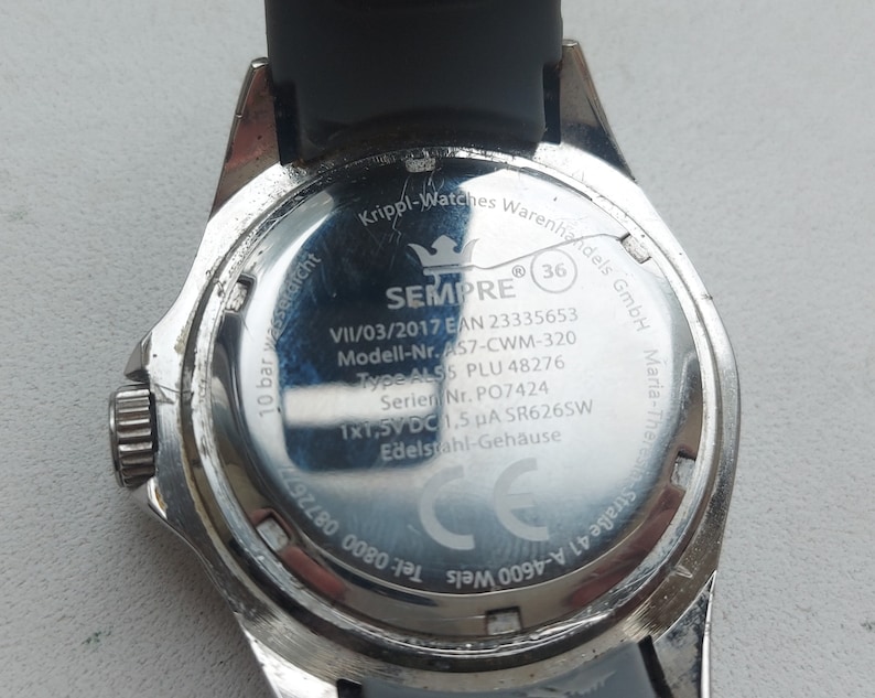 May include: The back of a silver watch with a black band. The watch face has the text "SEMPRE" and "10 bar wasserdicht" printed on it. The watch also has the text "VII/03/2017 EAN 23335653 Modell-Nr. AS7-CWM-320 Type ALS5 PLU 48276 Serier Nr. PO7424 1x1,5V DC 1,5 &mu;&Alpha; SR626SW Edelstahl-Geh&auml;use" printed on it.