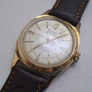 May include: Vintage gold-toned Selecta Deluxe 21 Jewel wristwatch with a cream-colored face and black leather band. The watch features gold-colored hands and hour markers, with the words "Shockproof, Waterproof, Antimagnetic" on the face.