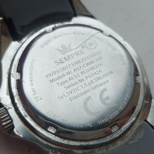 May include: The back of a silver watch with a black rubber band. The watch face is engraved with the words "SEMPRE", "VII/03/2017", "EAN 23335653", "Modell-Nr. AS7-CWM-320", "Type AL55 PLU 48276", "Serien Nr. PO7424", "1x1,5V DC 1,5 &mu;&Alpha; SR626SW", "Edelstahl-Geh&auml;use", "GmbH Maria-." and a CE mark.