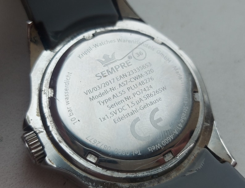 May include: The back of a silver watch with a black rubber band. The watch face is engraved with the words "SEMPRE", "VII/03/2017", "EAN 23335653", "Modell-Nr. AS7-CWM-320", "Type AL55 PLU 48276", "Serien Nr. PO7424", "1x1,5V DC 1,5 &mu;&Alpha; SR626SW", "Edelstahl-Geh&auml;use", "GmbH Maria-." and a CE mark.