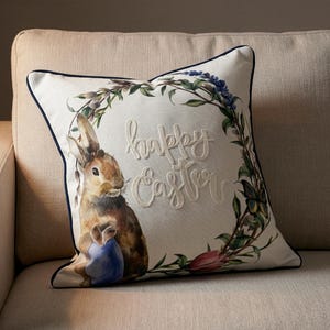 May include: Square decorative pillow with a watercolor illustration of a brown bunny holding a blue egg. The pillow has a white background with a navy blue trim and a floral wreath. The words "happy Easter" are embroidered in white.