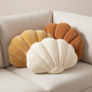 May include: Three seashell-shaped decorative pillows in shades of yellow, cream, and brown rest on a light-colored sofa. The pillows have a soft, velvety texture and are arranged in a visually appealing display.