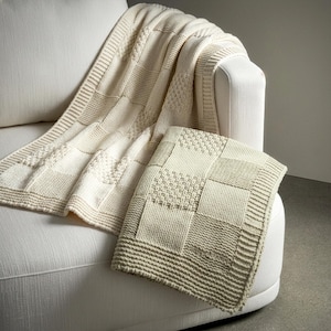 May include: Cream-colored knitted throw blanket and matching pillow. The blanket is draped over the arm of a white sofa, with the pillow resting on the sofa. The blanket and pillow feature a patchwork design with various textured knit patterns.