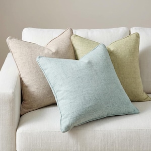 May include: Four decorative throw pillows in neutral colors rest on a white sofa. The pillows are in shades of beige, light green, and blue. The pillows have a textured appearance and are arranged in a casual, inviting display.