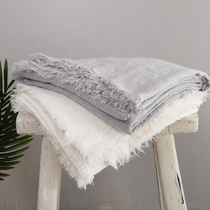 May include: Two fringed throw blankets, one white and one gray, are folded and draped over a white wooden stool.