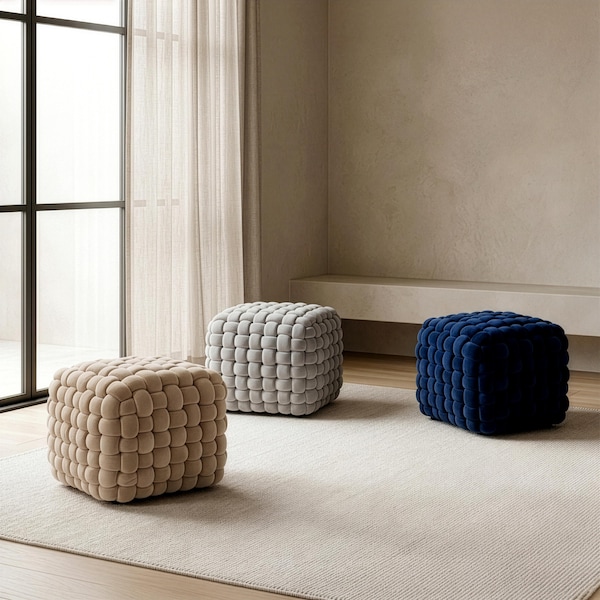 May include: Three cube-shaped ottomans in a neutral-toned room. One is beige, one is gray, and the third is a deep blue. Each ottoman has a woven, textured appearance, and they sit on a light-colored rug.