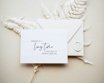 May include: A white card with black text that reads "FOREVER IS A LONG TIME I CAN'T WAIT TO SPEND IT WITH YOU". The card is sitting on a light brown surface with a white envelope and white fern branches.