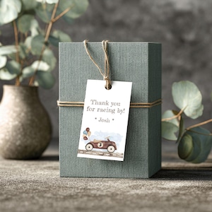 Può includere: Una scatola regalo verde acqua legata con spago, con un'etichetta bianca che recita "Thank you for racing by!" e "* Josh *." L'etichetta presenta un'illustrazione ad acquerello di un'auto da corsa marrone con palloncini. Un vaso con eucalipto è sullo sfondo.