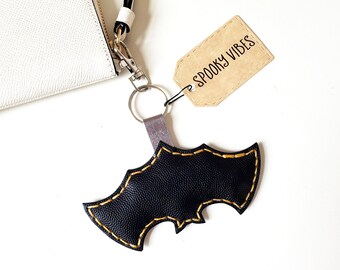 May include: Black bat-shaped keychain with gold stitching and a tag that reads "Spooky Vibes." The keychain is attached to a silver keyring and a gray strap. A white textured clutch is in the background.