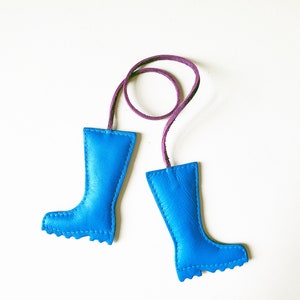 May include: A pair of bright blue leather boot-shaped keychains with a purple cord connecting them. The boots have a stitched design and a textured sole. The keychains are set against a white background.