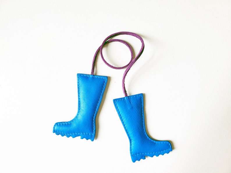 May include: A pair of bright blue leather boot-shaped keychains with a purple cord connecting them. The boots have a stitched design and a textured sole. The keychains are set against a white background.