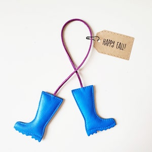 May include: A whimsical fall-themed ornament featuring two bright blue rain boots connected by a purple cord. A small, brown tag with the words "HAPPY FALL!" is attached. The boots are made of a smooth material.