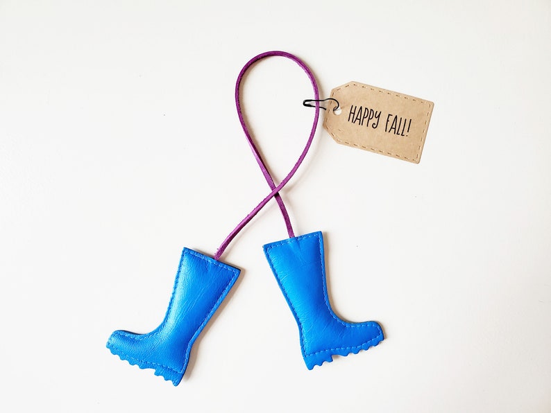 May include: A whimsical fall-themed ornament featuring two bright blue rain boots connected by a purple cord. A small, brown tag with the words "HAPPY FALL!" is attached. The boots are made of a smooth material.