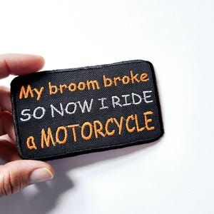 May include: A rectangular black embroidered patch with a stitched border. The text reads My broom broke SO NOW I RIDE a MOTORCYCLE in orange and white thread. The patch features a textured fabric background and is held against a plain white surface.