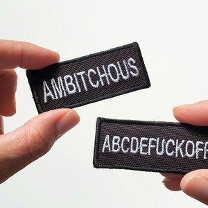 May include: Two black rectangular patches with white embroidered text are held up against a white background. One patch reads "AMBITCHOUS" and the other reads "ABCDEFUCKOFF". The patches have a black border.