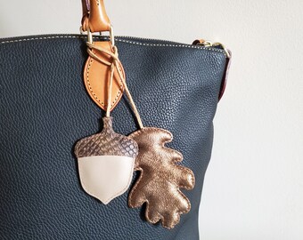May include: A dark blue leather handbag with a brown leather handle and two leather charms. One charm is an acorn with a brown and white design, and the other is a gold leaf.