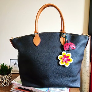 May include: A black leather tote bag with brown leather handles and a zipper closure. The bag has a ladybug and flower charm attached to it.