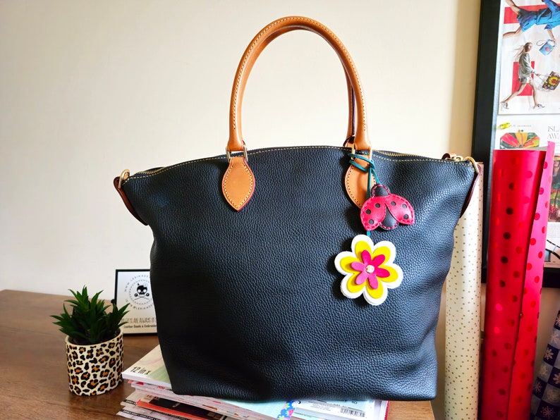 May include: A black leather tote bag with brown leather handles and a zipper closure. The bag has a ladybug and flower charm attached to it.
