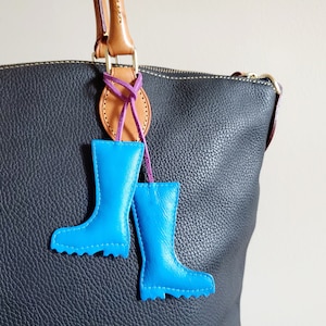 May include: A pair of bright blue leather boot charms hanging from a brown and purple leather accent on a black leather handbag. The charms are detailed with stitching and a scalloped edge.
