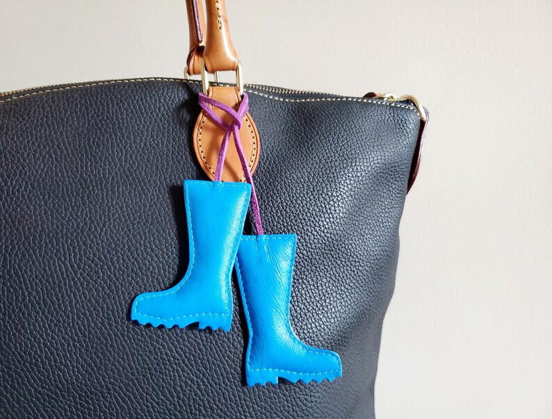 May include: A pair of bright blue leather boot charms hanging from a brown and purple leather accent on a black leather handbag. The charms are detailed with stitching and a scalloped edge.