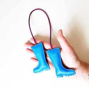 May include: A pair of small, blue leather boot-shaped ornaments with a purple cord. The boots have a textured appearance and are stitched around the edges. The cord is attached to the top of each boot.
