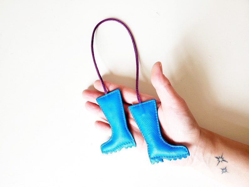 May include: A pair of small, blue leather boot-shaped ornaments with a purple cord. The boots have a textured appearance and are stitched around the edges. The cord is attached to the top of each boot.