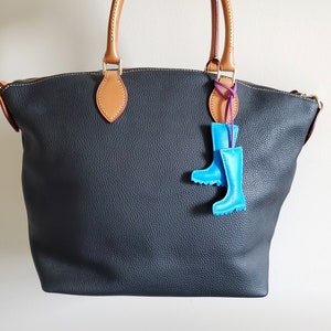 May include: A dark gray leather tote bag with tan handles and accents. A pair of light blue rain boot charms hang from the handle. The bag has a zipper closure and a textured surface.
