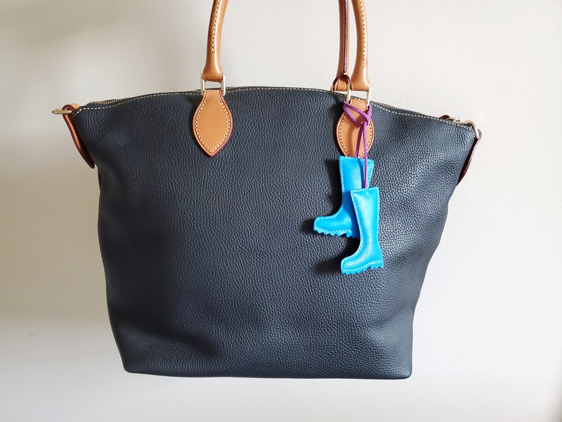 May include: A dark gray leather tote bag with tan handles and accents. A pair of light blue rain boot charms hang from the handle. The bag has a zipper closure and a textured surface.