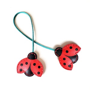May include: Two red ladybug-shaped leather bookmarks with black spots and black wings, connected by a teal ribbon.