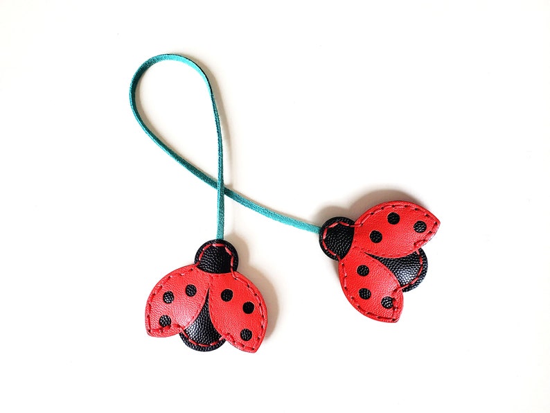 May include: Two red ladybug-shaped leather bookmarks with black spots and black wings, connected by a teal ribbon.
