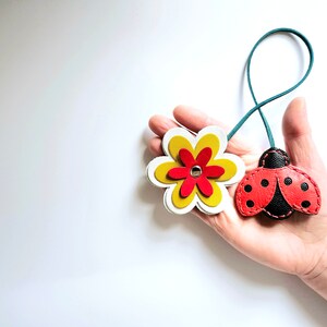 May include: A leather keychain with a red and black ladybug charm and a yellow and white flower charm. The charms are attached to a teal cord.