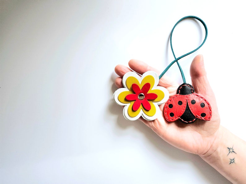 May include: A leather keychain with a red and black ladybug charm and a yellow and white flower charm. The charms are attached to a teal cord.
