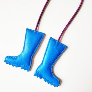 May include: Two blue leather boot-shaped ornaments with purple cords for hanging. The boots have a stitched design and a scalloped edge at the bottom. The background is plain white.