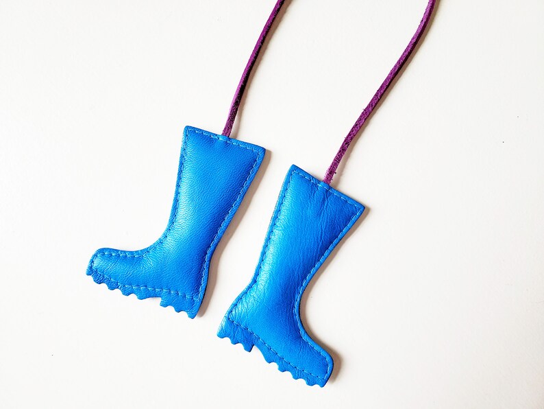 May include: Two blue leather boot-shaped ornaments with purple cords for hanging. The boots have a stitched design and a scalloped edge at the bottom. The background is plain white.