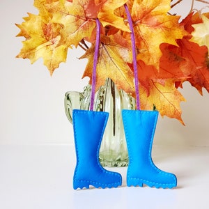 May include: Two bright blue leather boot ornaments with white stitching hang from purple ribbons. The boots are displayed in front of a vase with fall-colored leaves, creating a seasonal decorative theme.