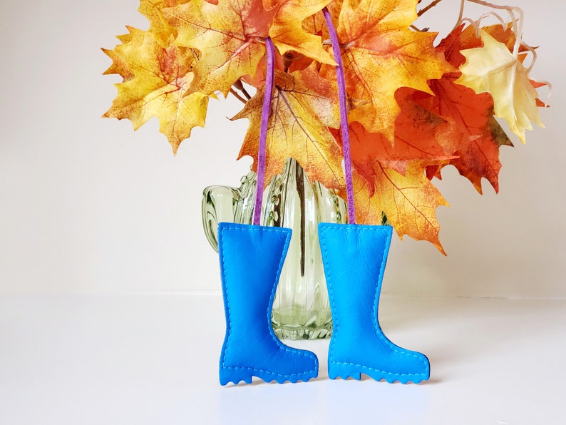 May include: Two bright blue leather boot ornaments with white stitching hang from purple ribbons. The boots are displayed in front of a vase with fall-colored leaves, creating a seasonal decorative theme.