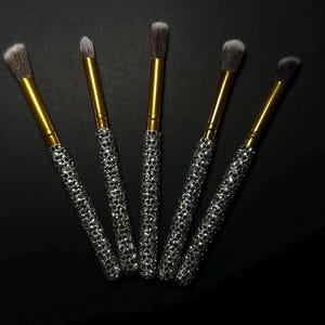 May include: Five makeup brushes with gold handles and silver rhinestone-covered handles. The brush heads are various shapes and sizes, with soft, dark bristles. The brushes are arranged on a black background.